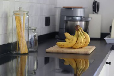 Siyah bir mutfak tezgahının tahta kesme tahtasının üzerinde duran bir grup sarı muzun portresi. Lezzetli enerji veren yemekler yemeğe hazır ve spordan önce ideal..