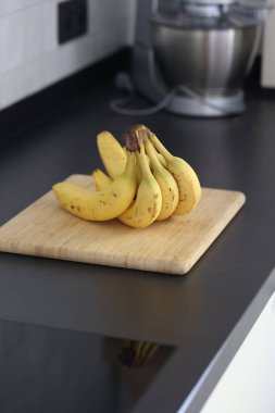 Siyah bir mutfak tezgahının tahta kesme tahtasının üzerinde duran bir grup sarı muzun portresi. Lezzetli enerji veren yemekler yemeğe hazır ve spordan önce ideal..