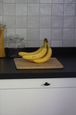 Siyah bir mutfak tezgahının tahta kesme tahtasının üzerinde duran bir grup sarı muzun portresi. Lezzetli enerji veren yemekler yemeğe hazır ve spordan önce ideal..