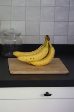 Siyah bir mutfak tezgahının tahta kesme tahtasının üzerinde duran bir grup sarı muzun portresi. Lezzetli enerji veren yemekler yemeğe hazır ve spordan önce ideal..