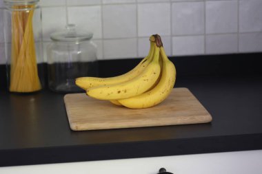 Siyah bir mutfak tezgahının tahta kesme tahtasının üzerinde duran bir grup sarı muzun portresi. Lezzetli enerji veren yemekler yemeğe hazır ve spordan önce ideal..