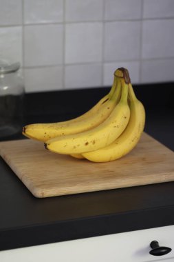 Siyah bir mutfak tezgahının tahta kesme tahtasının üzerinde duran bir grup sarı muzun portresi. Lezzetli enerji veren yemekler yemeğe hazır ve spordan önce ideal..