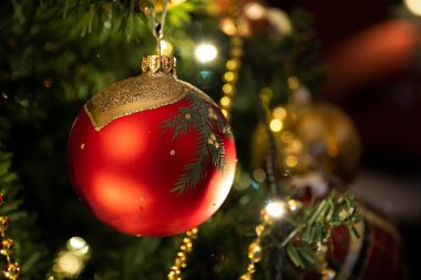 Parlak ışıkları ve yeşil yaprakları olan, bayram sıcaklığını ve şenlikli tezahüratı çağrıştıran güzel dekore edilmiş bir ağaca asılı klasik kırmızı Noel süsü..