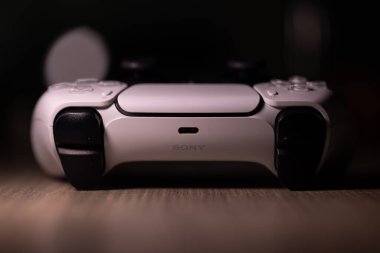 Brecht, Belçika - 18 Nisan 2025: Orjinal beyaz sony playstation 5 kablosuz kontrol cihazının ahşap bir masanın üzerinde duran koyu renkli bir portresi. Bir PS5 video oyunu oynamaya hazır. 