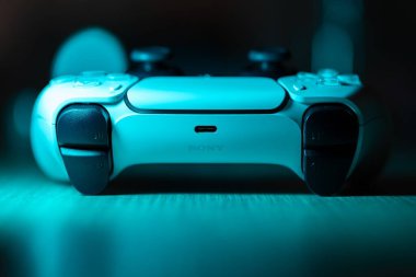 Brecht, Belçika - 18 Nisan 2025: Orjinal beyaz sony playstation 5 kablosuz kontrol cihazının ahşap bir masanın üzerinde duran mavi neon bir portresi. Bir PS5 konsol oyunu oynamaya hazır.