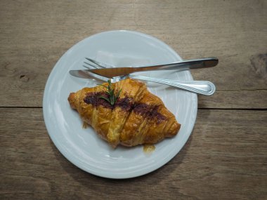 Kruvasan beyaz bir tabakta. Bıçak ve çatal ahşap arka planda