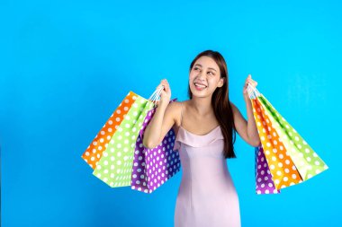 Mutlu gülümseyen Asyalı bayan alışverişçi elinde alışveriş torbalarıyla yürürken kameraya bakıyor. Modaya uygun giyinen kız mavi arka planda izole edilmiş. Stüdyo çekiminde vücut uzunluğunun yarısı.