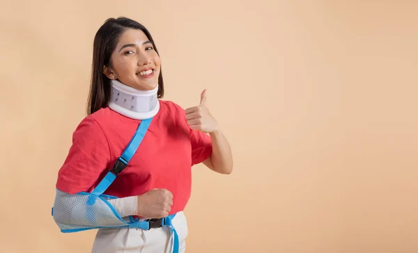 Beautiful young asian woman with broken arm in soft splint suffering a sore arm showing thumbs up sign isolated on beige background, accident insurance concept.