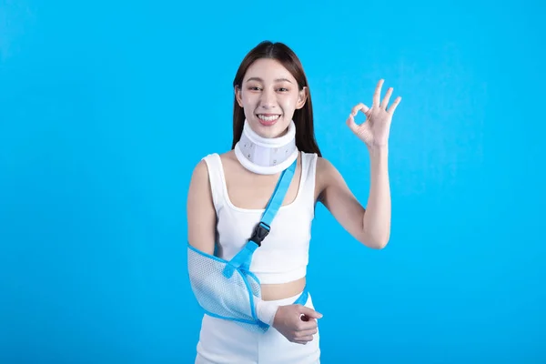 Beautiful young asian woman with broken arm in soft splint suffering a sore arm showing okay sign isolated on blue background, accident insurance concept.