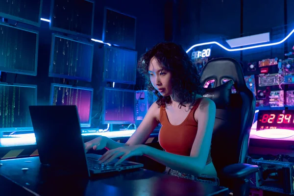 Female Asian hacker in hideout place with dark environment and multiple displays and cables, trying to break into data servers and infect the system with a virus.