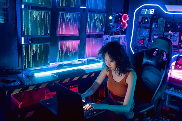 Female Asian hacker in hideout place with dark environment and multiple displays and cables, trying to break into data servers and infect the system with a virus.