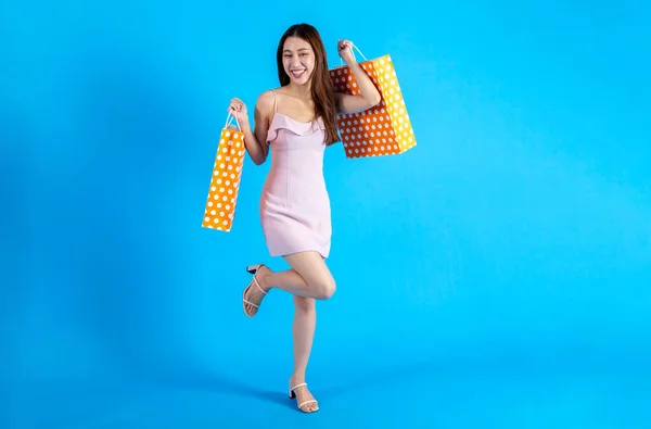 Happy smiling Asian female shopper holding a bunch of shopping bags while walking and looking at camera. Fashionable girl in dress isolated on blue color background. Half body length in studio shot.
