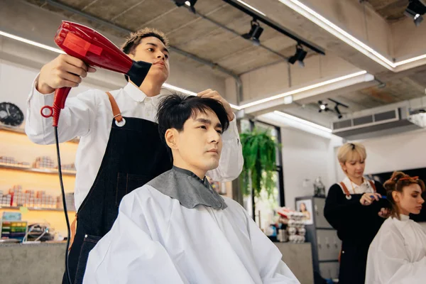 現代的な理髪店でハンサムな若い男性の髪を乾燥エプロンを身に着けている若い男性アジアの美容師は仕事に集中しながら — ストック写真