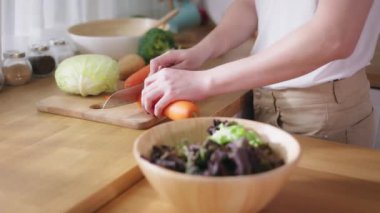Close-up of young man in white t-shirt slicing and cutting raw vegetables such as cabbage and carrot in kitchen at home. Vegetarian making a salad.