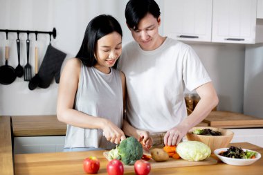 Asian couple in love relationship making a healthy salad together in the kitchen. Smiling happy boyfriend and girlfriend enjoying cooking activity at home apartment.