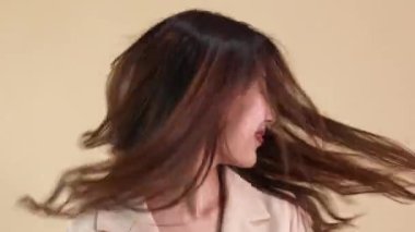 Genç Asyalı mutlu, günlük iş elbisesi içinde gülümseyen bir kadın. Bej arka planda uçuşan saçlar var.