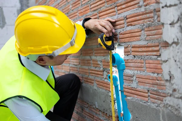 Young Asian engineer in engineering uniform and helmet at construction site using a measurement tape to measure for precision. 30s professional foreman working on structure building at workplace