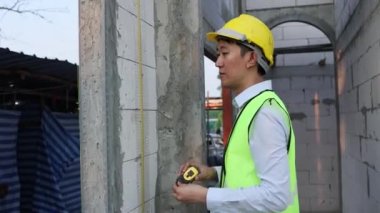 İnşaat alanında mühendis üniforması ve miğferi olan genç Asyalı bir mühendis kesinliği ölçmek için mezura kullanıyor. 30 'ların profesyonel ustabaşı işyerindeki yapı inşaatında çalışıyor.
