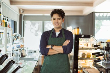 Önlüklü genç Asyalı erkek barista kameraya bakarken kollarını kavuşturup gülümsüyor.