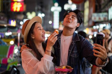 Bangkok 'taki Yaowarat Yolu' ndaki kalabalıkla gece pazarında sokak tezgahından yemek yerken selfie çeken ve video blogu kaydeden Asyalı çift turist.