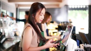Asyalı kafe çalışanı bir kafede tablet bilgisayar kullanırken cep telefonuyla yapılan ön siparişi kabul ediyor. Kadın garson cep telefonuyla konuşuyor. Ev yemeği teslimatı kavramı.