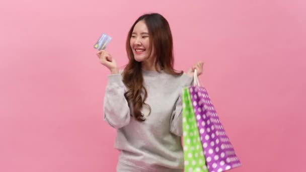 ピンクのバックグラウンドで買い物バッグを持っている間 クレジットカードを持っている幸せなアジアの若い女性の買い物客の肖像画 オンライン決済コンセプトでオンラインショッピング — ストック動画