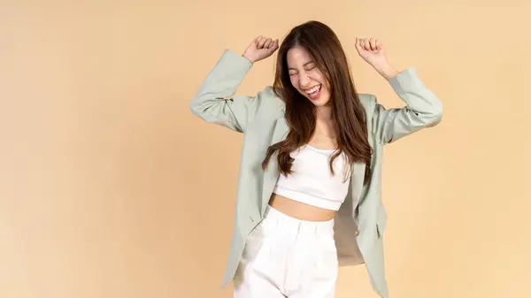 Young Asian happy smiling woman in casual business suit raising her hands up like a winner isolated over beige background - with empty copy space