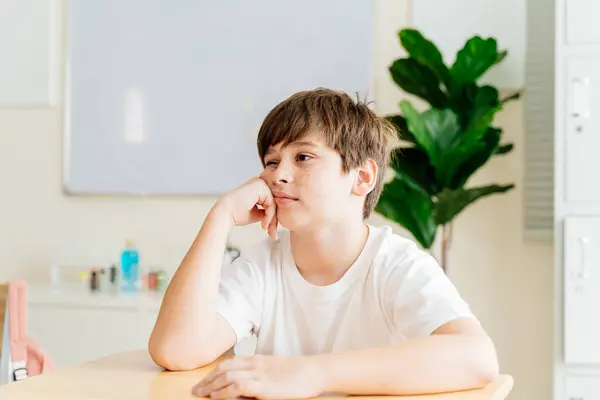 stock image Freckled boy in classroom setting, thinking and daydreaming. Young student in white shirt at desk, casual and relaxed, showcasing youth and innocence. Education and learning environment portrait