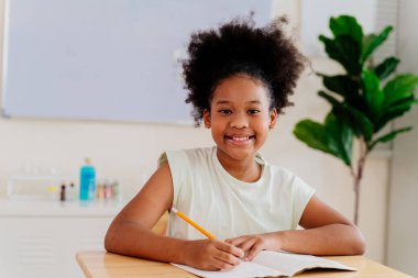 Sınıfta yazı yazarken gülümseyen mutlu Afrikalı Amerikalı kız. Afro saçlı genç bir öğrenci, elinde kalem ve defterle bir masada oturuyor.