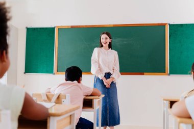 Genç Kafkas öğretmeni çeşitli anaokulu öğrencilerini parlak sınıflara çekiyor. Gülümseyen eğitimci yeşil karatahtanın önünde güvenle duruyor, erken öğrenme ortamını teşvik ediyor..