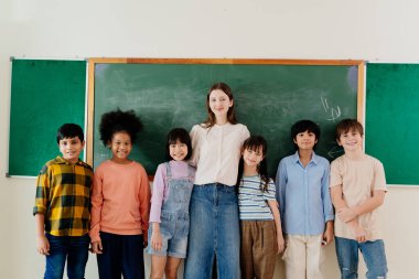 Çeşitli ilkokul öğrencileri sınıf tahtası önünde öğretmenle poz veriyor. Çok kültürlü çocuklar ve eğitimciler ilkokul ortamında kapsayıcı öğrenme ortamı sergilediler