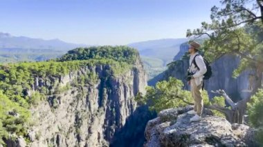 Beyaz tişörtlü, kahverengi şapkalı, sırtında sırt çantası olan beyaz bir adam elinde kamerayla ormanın etrafındaki büyük kanyonun önündeki uçurumun kenarına gelir ve panoramik manzaraya hayran kalır.