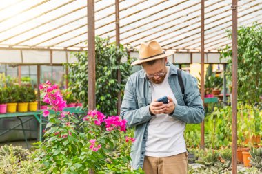 Akıllı telefon kullanan şapkalı genç bahçıvan siparişleri alır ve seradaki müşterilerin siparişlerini işlemek için bitkilerin fotoğraflarını çeker. Bahçe merkezinde süs bitkileri, bitkiler satılıyor.