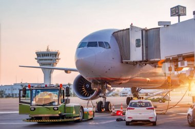 Aircraft is being serviced by airfield ground services at gangway of terminal airport building, preparing for towing and launching into flight in the evening at sunset clipart