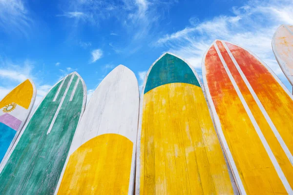 Several surfboards of different colorful saturated yellow, green, red colors lined up against the background of a blue summer sky with clouds at sunny day
