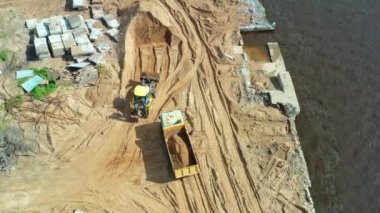Tractor with a bucket in front loads the soil sand into the truck body at the construction site of the supports of the embankment of the river, bay, sea. Aerial top view