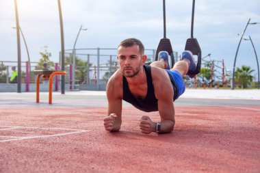 Spor sahasındaki adam tahta bir pozisyonda duruyor, basın üzerinde bir egzersiz, ellerinin dirseklerine yaslanıyor, halat halkalarının arkasındaki bacaklar.