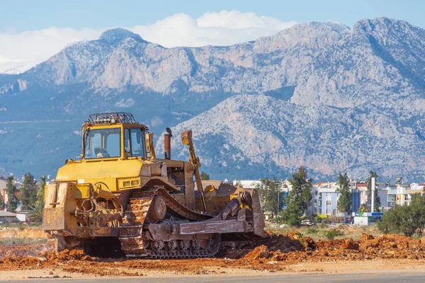 Buldozer, ıssız bir arazinin kentsel alanındaki zemini dağların arka planına karşı düzleştiriyor.