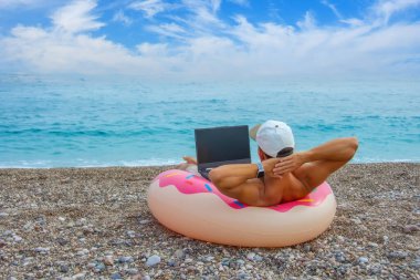 Şapkalı bir adam, şişirilmiş bir çemberin içinde kumsalda pembe kremalı bir çörek şeklinde oturur ve internette dizüstü bilgisayarda çalışır. Tatilde istihdam kavramı, iş ve boş zamanı birleştirmek.