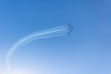Mavi gökyüzünde akrobatik uçuş yapan bir uçağın silüeti. Arkasında duman izi var.