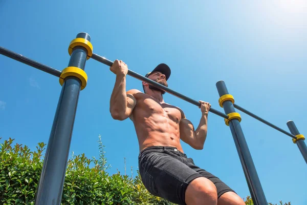 stock image Muscular man in a cap doing reverse pull-up on the horizontal bar in the park outdoors