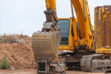 Çöp dolu bir çöp sahasında sarı kazıcı yüklemeye hazır bir kamyonun yanında.