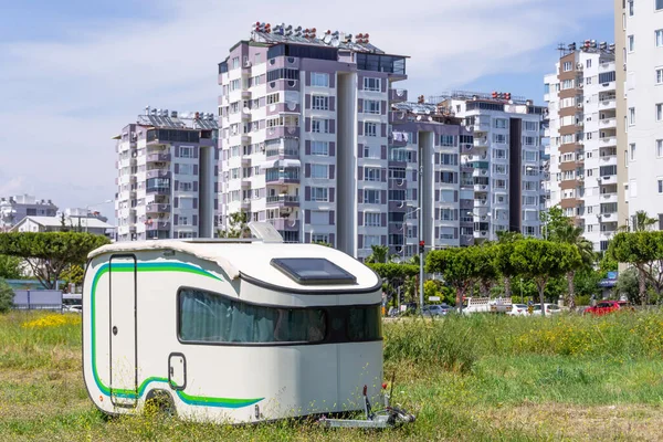 Mobile home trailer on wheels van parked in the street on the meadow of the city park, against the background of multi-storey residential buildings.