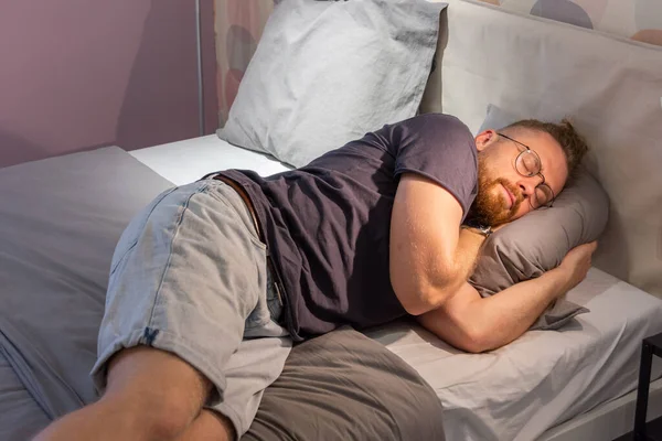 Young guy man beard fell asleep in glasses on a bed in clothes in the bedroom with the lights on. Fatigue, lack of sleep, stress, comfort, randomness