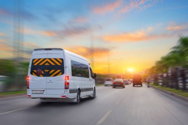 Tanımlanamayan küçük beyaz yolcu minibüsü şehir trafiğinde otoyolda acele ediyor, arka planda gündoğumu gökyüzü. Charter hızlı otobüs minibüsü hareket bulanıklığı etkisi ile acele.