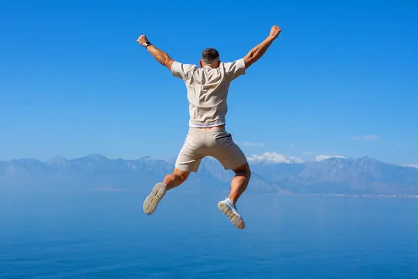 Молодой Человек Прыгает Скале Поднятыми Руками Голубые Вершины Гор Океана — стоковое фото