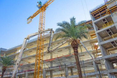 Sarı uzun vinç, inşaat halindeki uzun apartmanın beton iskeleti, yakınlardaki palmiye ağacı..