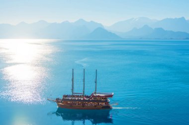Yelkenli tarihi ahşap gemi, deniz yüzeyinin sularında yüzüyor. Dağ zirveleriyle çevrili, parlak güneşli, gökyüzü manzaralı bir koy.