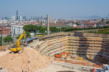 Toprakları kazılmış bir metro istasyonunun çukuru. Çalışan kazıcılar toprağı kazıp çıkarıyorlar. Arka planda bir şehir, evler, binalar, yerleşim alanı var.