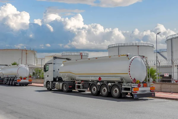 Trucks with trailers of white fuel tanks are parked next to a huge oil storage barrel